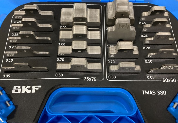 Obraz jest profesjonalną i kompletną prezentacją produktu, ukazującą zestaw SKF TMAS 380 jako wszechstronne i niezbędne narzędzie dla każdego profesjonalnego działu utrzymania ruchu. Kompozycja jest czysta, zorganizowana i techniczna. Dominują w niej lśniące metaliczne odcienie stali nierdzewnej podkładek, kt&oacute;re kontrastują z ciemną, wytrzymałą walizką. Całość emanuje precyzją, wszechstronnością i gotowością do działania.  Pierwszy plan &ndash; Kompletne rozwiązanie do osiowania:  W centrum uwagi znajduje się solidna, otwarta walizka transportowa, kt&oacute;ra w pełni eksponuje bogate i uporządkowane wnętrze zestawu. Kluczowe elementy to:  Wytrzymała walizka: Ciemna, przemysłowa walizka z wyraźnie widocznym białym logo "SKF" oraz oznaczeniem modelu "TMAS 380". Jej otwarta forma zaprasza do zapoznania się z imponującą zawartością.  Bogaty zestaw podkładek: Wnętrze walizki, wyłożone specjalnie wyprofilowaną pianką lub przegr&oacute;dkami, zawiera imponującą liczbę 380 podkładek precyzyjnych w dw&oacute;ch najpopularniejszych rozmiarach:  50 x 50 mm: Do mniejszych maszyn i precyzyjnych regulacji.  75 x 75 mm: Uniwersalny rozmiar do większości standardowych silnik&oacute;w i pomp.  Perfekcyjna organizacja: Każdy rozmiar podkładki ma swoją dedykowaną, wyraźnie oznaczoną sekcję, a w niej poszczeg&oacute;lne stosy są uporządkowane według dziewięciu r&oacute;żnych grubości (od 0,05 mm do 2,00 mm). Taki układ wizualnie komunikuje łatwość i szybkość doboru odpowiedniej podkładki w każdych warunkach.  Najwyższa jakość wykonania: Podkładki, wykonane z wysokogatunkowej stali nierdzewnej, lśnią w studyjnym oświetleniu. Na każdej z nich widoczne jest precyzyjne, laserowo grawerowane oznaczenie grubości i wymiaru, co eliminuje pomyłki. Charakterystyczne wycięcie ułatwiające montaż jest dobrze widoczne na każdym stosie.  Tło i branding:  Tło jest neutralne (np. białe lub jasnoszare), aby nie odwracać uwagi od bogactwa zestawu. Gł&oacute;wne elementy brandingowe to logo SKF na walizce oraz oznaczenia na samych podkładkach, co jest gwarancją autentyczności i najwyższej jakości.  Przesłanie i hasło reklamowe (optymalizacja SEO):  Obraz niesie jasny komunikat o wszechstronności, precyzji i profesjonalizmie. Skierowany jest do technik&oacute;w utrzymania ruchu, firm serwisowych i inżynier&oacute;w, kt&oacute;rzy potrzebują jednego, kompletnego zestawu do osiowania maszyn o r&oacute;żnych gabarytach, pokrywającego większość standardowych zastosowań przemysłowych.  Całość może być wzmocniona hasłem, kt&oacute;re zawiera kluczowe frazy:  "Jeden zestaw, pełna wszechstronność. Podkładki do osiowania SKF TMAS 380."  "Wszechstronność i precyzja w jednej walizce. SKF TMAS 380 dla profesjonalist&oacute;w."  "Twoje kompletne rozwiązanie do osiowania maszyn &ndash; Zestaw SKF TMAS 380."
